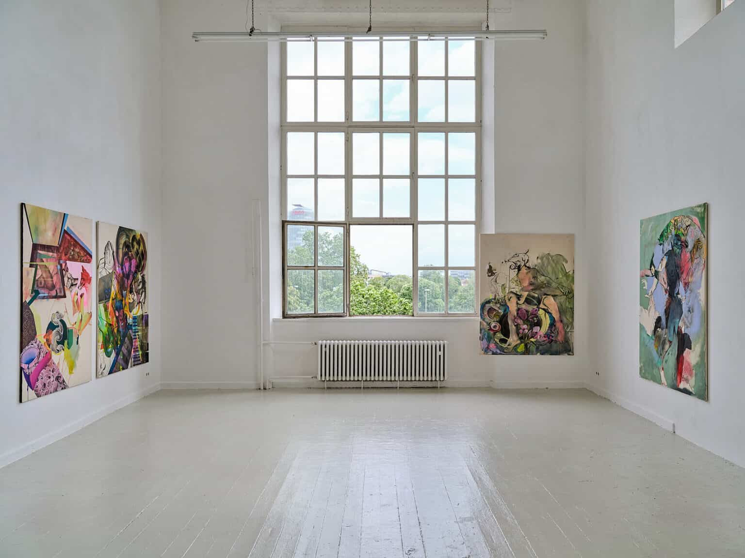 Foto: Ansicht eines Raumes mit weißen Wänden, grauem Dielenboden und einem großen Fenster. Im Raum hängen großformatige Leinwandarbeiten auf ähnlicher Höhe. Eine der Arbeiten Ragt über einen Vorsprung an der Wand in die Einbuchtung des Fensters hinein.