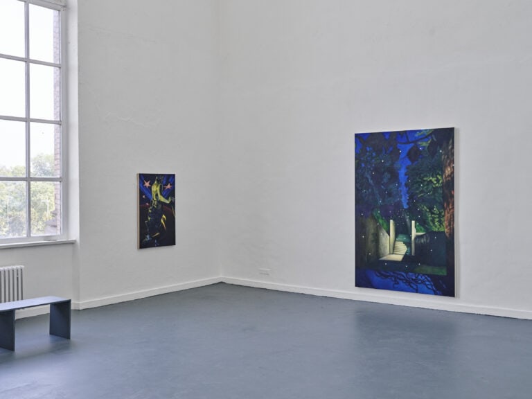 Foto: Aufnahme eines Ausstellungsraums mit weißen Wänden, grauem Boden und einem großen Fenster. An den Wänden hängen zwei unterschiedlich große Gemälde. Eine Bank steht vor dem Fenster.