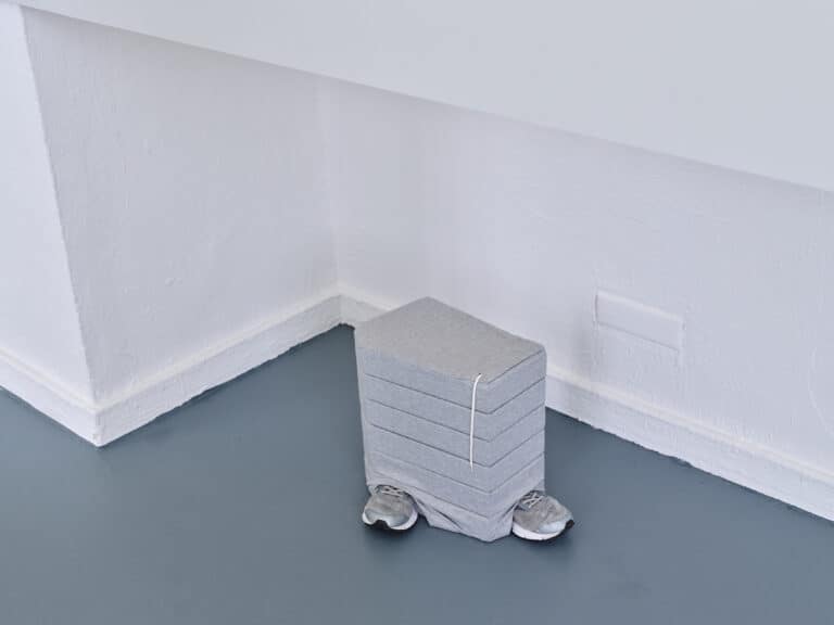 Foto: Detailaufnahme eines Kunstwerks. Stapelartige Säule mit Stoffbezogen aus der zwei Turnschuhe herausstehen.