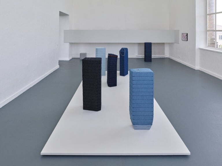 Foto: Aufnahme eines Ausstellungsraums mit weißen Wänden, grauem Boden und einem großen Fenster. In der Raummitte ist eine fünfteilige Kunstinstallation aus Jeansstoff auf einer weißen Bodenplatte aufgebaut.