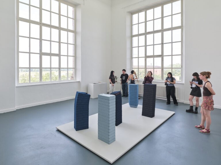 Foto: Aufnahme eines Ausstellungsraums mit weißen Wänden, grauem Boden und zwei großen Fenstern. In der Raummitte ist eine fünfteilige Kunstinstallation aus Jeansstoff auf einer weißen Bodenplatte aufgebaut. Mehrere Personen stehen im Raum und betrachten das Kunstwerk.
