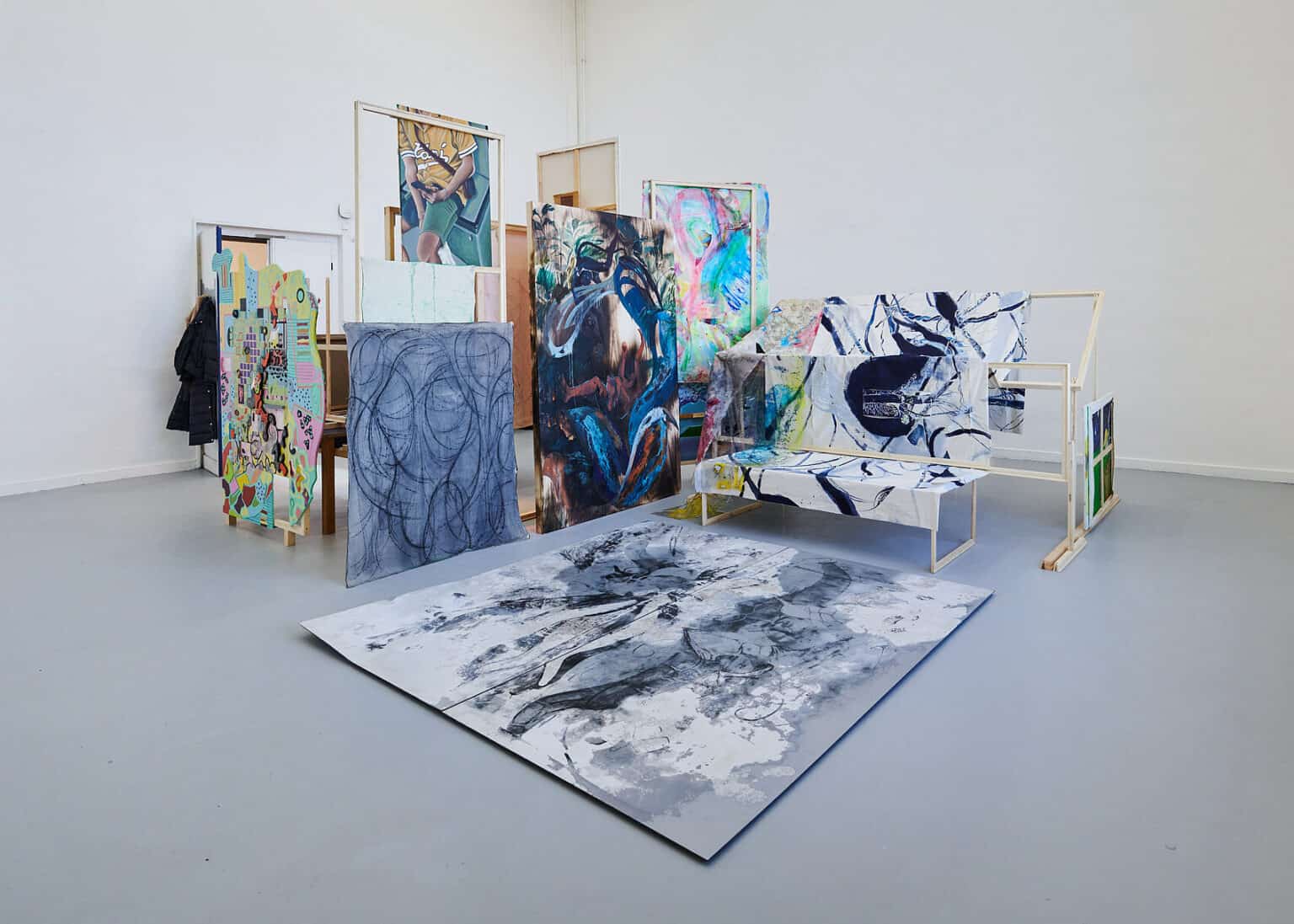 Foto: Ansicht eines Raumes mit weißen Wänden und grauem Boden. Im Raum befindet sich eine Konstruktion aus Holzlatten, an der Bilder und Textilien angebracht sind.