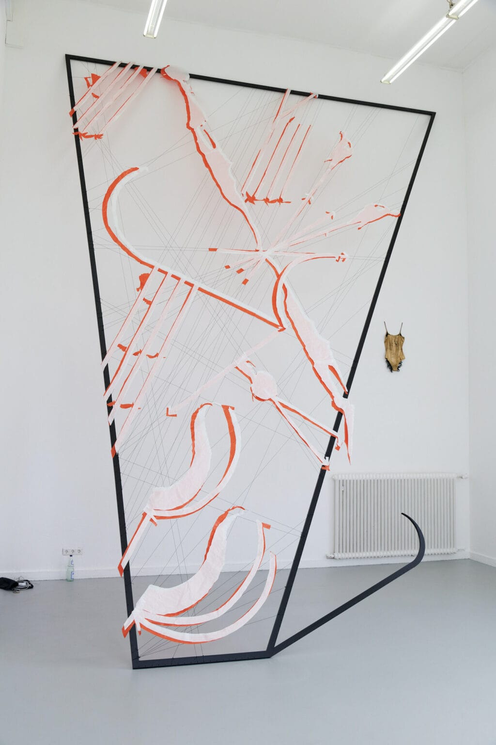 Foto: Ein Raum mit weißen Wänden, an denen ein Objekte hängt. Im Raum eine große Skulptur, die auf dem Boden steht und beinahe bis unter die Decke reicht.