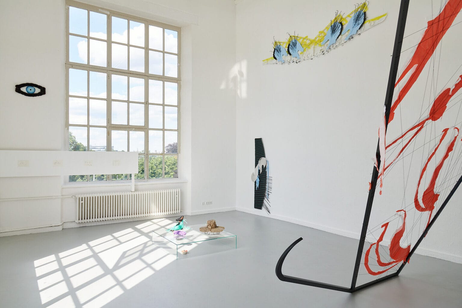 Foto: Ein Raum mit weißen Wänden, grauem Boden und einem großen Fenster. An den Wänden hängen auf unterschiedlicher Höhe Objekte. Im Raum stehen zwei Skulpturen. Vor dem Fenster und über die Wand hinweg erstreckt sich ein flaches Objekt.