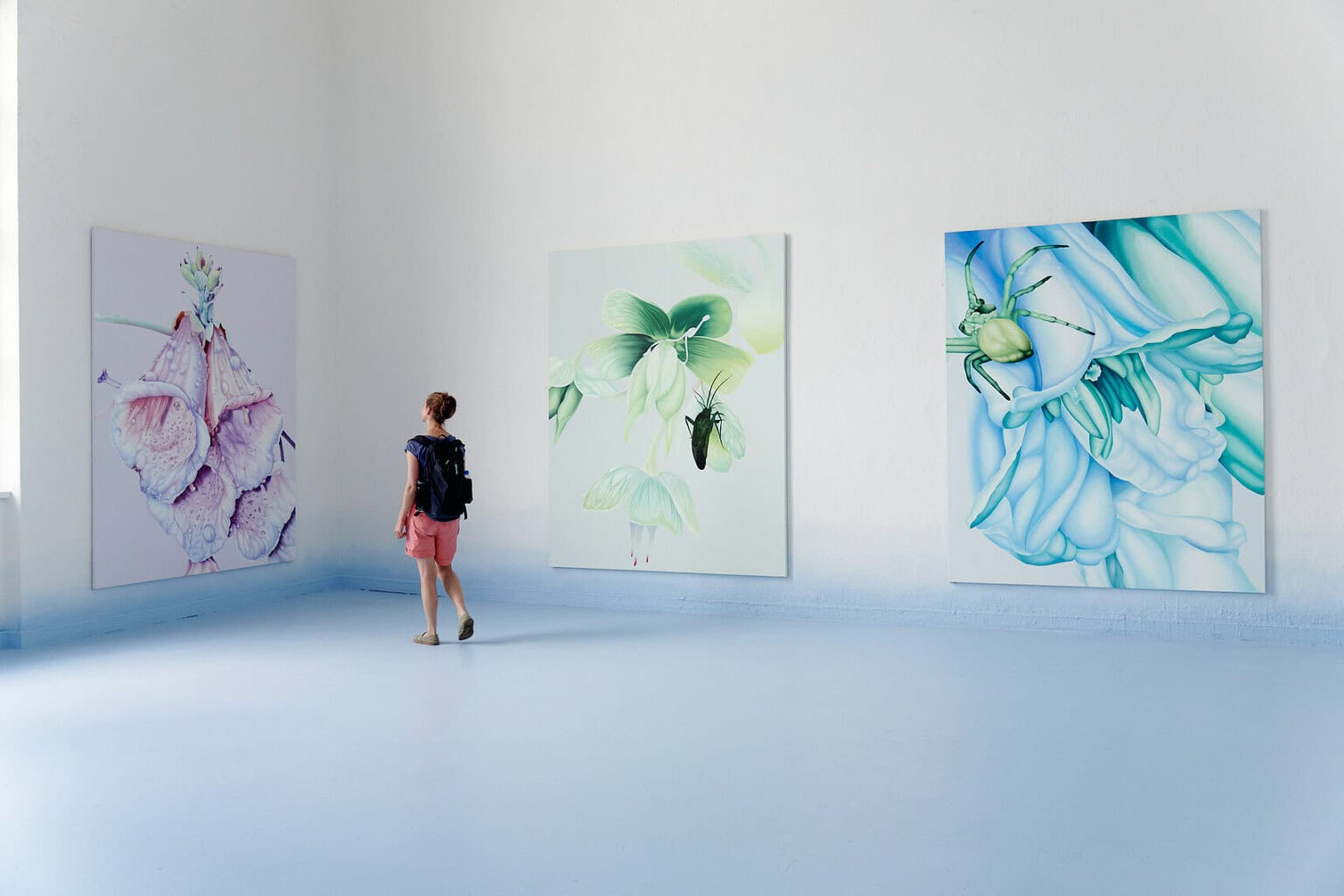 Foto: Ein Raum mit weißen Wänden und hellblauem Boden. Der untere Bereich der Wand ist ebenfalls mit hellblauer Farbe bemalt, die nach oben hin ausfaded. An der Wand hängen, auf gleicher Höhe, drei großformatige Leinwandarbeiten in ähnlichem Format. Durch den Raum bewegt sich eine Person.