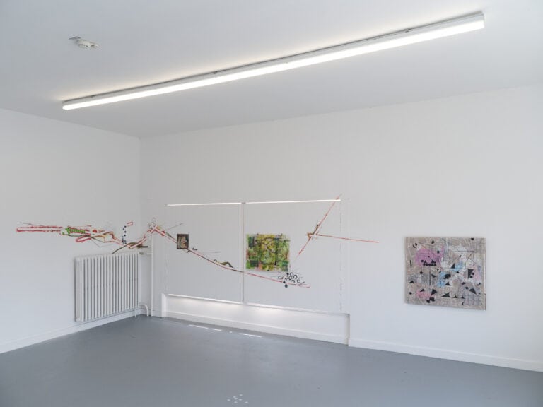 Foto: Aufnahme eines Ausstellungsraums mit weißen Wänden und grauem Boden, einem weißen Heizkörper und einer Lichtleiste an der Decke. An der Wand befinden sich mehrere abstrakte Gemälde, teilweise direkt auf die Wand aufgetragen, teilweise als Bilder.