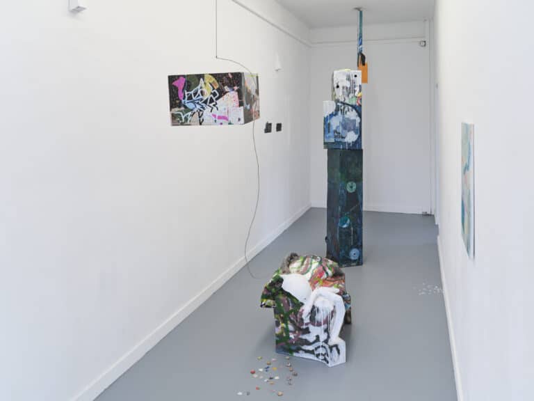 Foto: Aufnahme eines schmalen Ausstellungsraums mit weißen Wänden und grauem Boden. In der Mitte des Raums ist eine mehrteilige abstrakte Kunstinstallation aufgebaut. An den Wänden hängen einzelne Bilder.