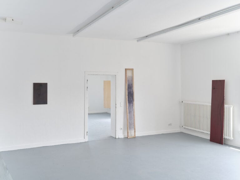 Foto: Aufnahme eines Ausstellungsraums mit weißen Wänden und grauem Boden. An den Wänden und dem Heizkörper sind schmale Farbflächengemälde angelehnt. Ein kleineres Gemälde hängt an der Wand.