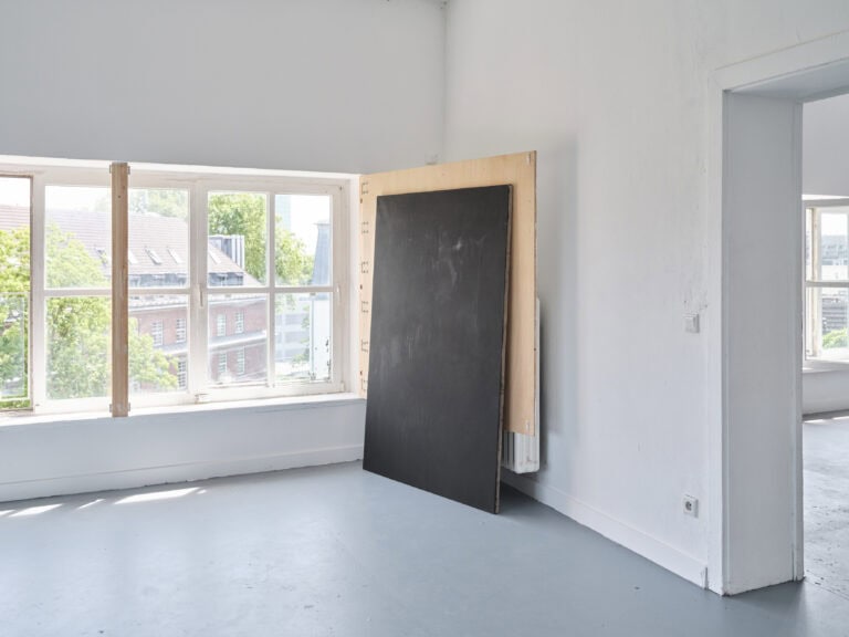Foto: Aufnahme eines Ausstellungsraums mit weißen Wänden, grauem Boden und Fensterflächen. In der Raumecke sind großformatige Farbflächengemälde angelehnt.