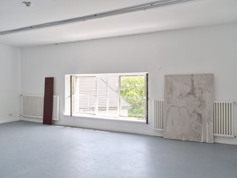 Foto: Aufnahme eines Ausstellungsraums mit weißen Wänden, grauem Boden und Fensterflächen. Neben dem Fenster sind großformatige Farbflächengemälde angelehnt.