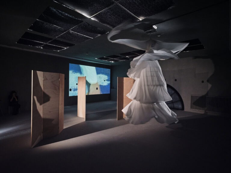 Foto: Aufnahme eines abgedunkelten Ausstellungsraums mit aufgestellten Holztrennwänden. An einer Wand ist ein Video projiziert. Weiße Textilien hängen drehend an der Decke.