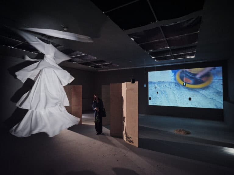 Foto: Aufnahme eines abgedunkelten Ausstellungsraums mit aufgestellten Holztrennwänden. An einer Wand ist ein Video projiziert. Weiße Textilien hängen drehend an der Decke.