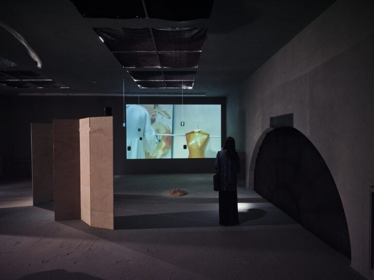 Foto: Aufnahme eines abgedunkelten Ausstellungsraums mit aufgestellten Holztrennwänden. An einer Wand ist ein Video projiziert. Eine Person steht im Raum und ist von hinten zu sehen.