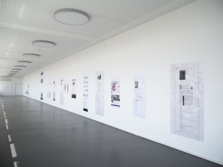 Foto: Aufnahme eines langen schmalen Ausstellungsraums mit weißen Wänden und grauem Boden. Entlang der rechten Wand sind Drucke aufgehängt.