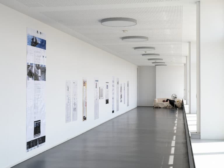 Foto: Aufnahme eines langen schmalen Ausstellungsraums mit weißen Wänden und grauem Boden, an dessen rechter Seite Fenster zu sehen sind. Entlang der linken Wand sind Drucke aufgehängt. Im rückwärtigen Bereich steht eine Couch. Eine Person liegt aus der Couch vor einem Ventilator.