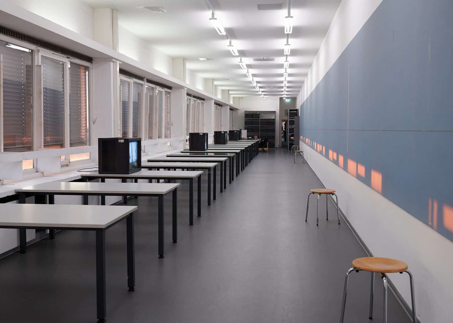 Foto: Ansicht eines länglichen Raumes mit weißen Wänden, grauem Boden und Fensterfront und heruntergelassenen Rollläden. Vor der Fensterfront steht eine Reihe aus Tischen. Auf mancher der Tische stehen eingeschaltete Fernseher. Über die rechte Wand des Raumes erstreckt sich ein blauer Streifen, auf den in kleinen Quadraten rötliches Sonnenlicht fällt. Vor der Wand stehen mehrere Hocker.