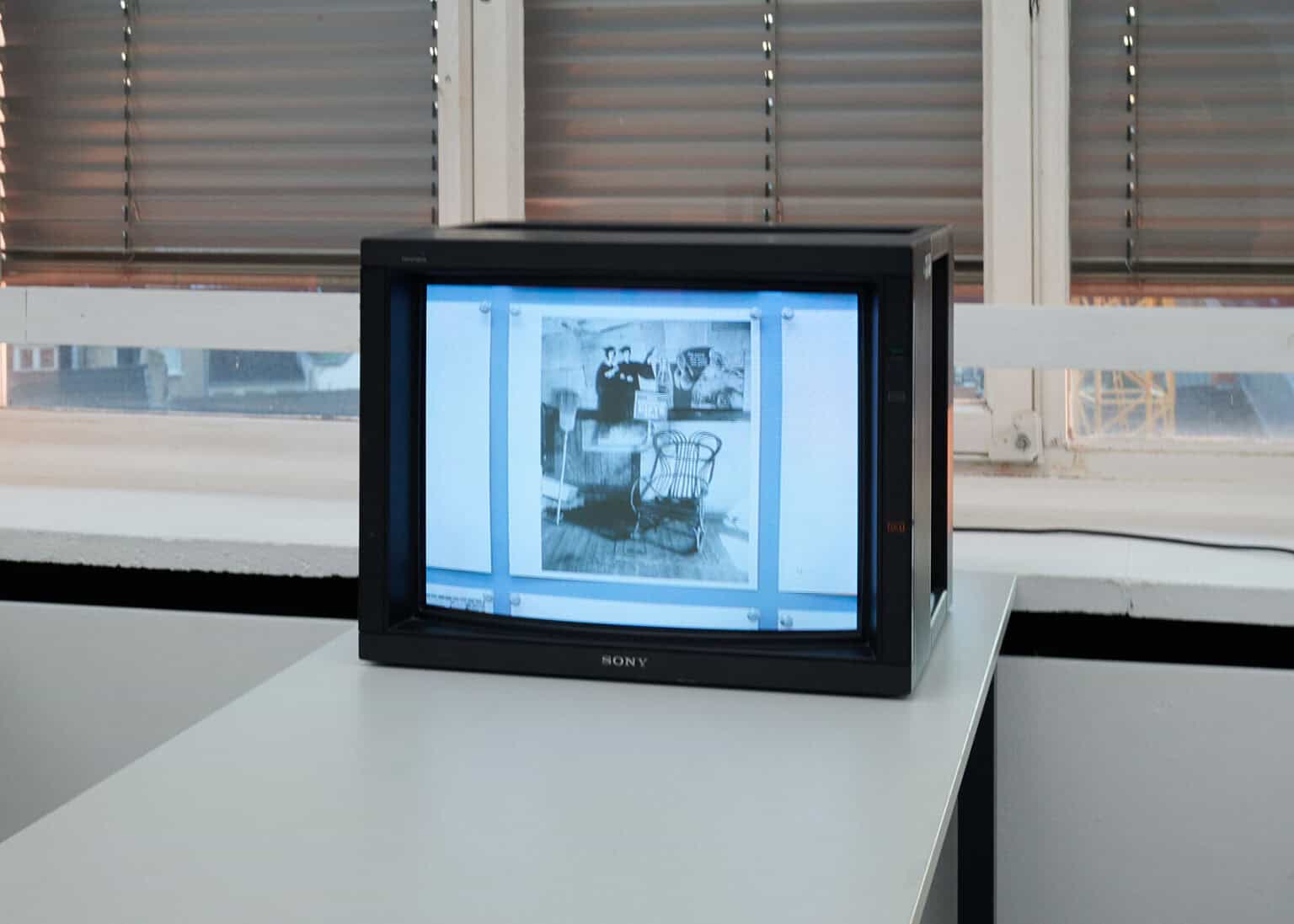 Foto: Nahaufnahme eines eingeschalteten Fernsehers auf einem Tisch vor einer Fensterfront mit herabgelassenen Rollläden.