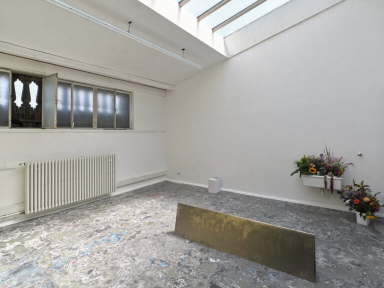 Foto: Aufnahme eines Ausstellungsraums mit weißen Wänden und einem gemusterten Boden. Eine abstrakte Kunstinstallation steht auf dem Boden. Im rückwärtigen Bereich ist ein Waschbecken zu sehen, in dem Blumen stehen. Weitere Blumen stehen in einem Eimer daneben.