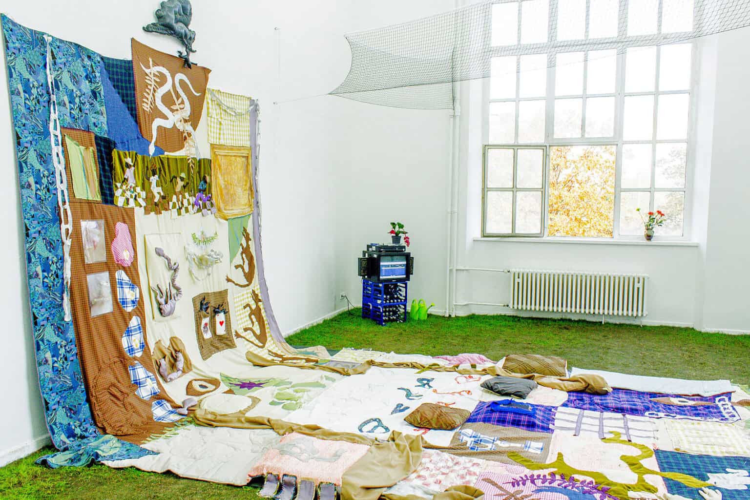 Foto: Ein Raum mit weißen Wänden und einem großen Fenster. Der Boden ist mit Rollrasen ausgelegt. An der Wand ist ein großes, farbiges, flickenartiges Textil befestigt, dass über die Wand herunter über den Boden reicht. Auf dem Textil liegen Kissen. In der Ecke des Raumes steht ein Fernseher auf zwei Getränkekästen. Daneben zwei Gießkannen. In der Oberen hälfte des Raumes ist zwischen den Wänden ein Netz aufgespannt.