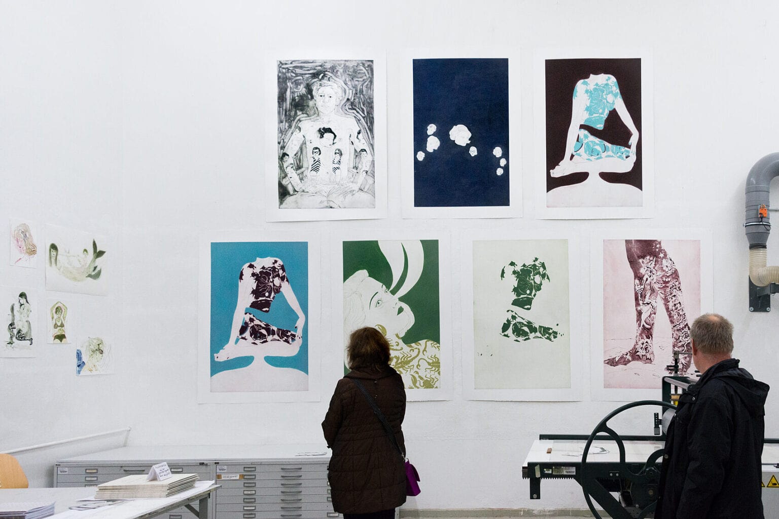 Foto: Ein Raum mit weißen Wänden. An der Wand des Raumes hängen bedruckte und bemalte Papiere im Klein- und Großformat über- und nebeneinander. Im Raum befinden sich außerdem Tische, eine Druckpresse und zwei Planschränke sowie zwei Menschen.