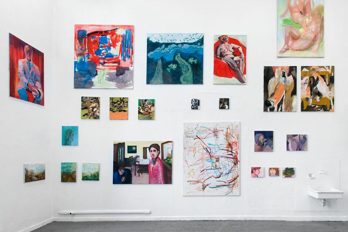 Foto: Ein Raum mit weißen Wänden und grauem Dielenboden. An den Wänden hängen mehrere Leinwände in unterschiedlichen Formaten neben- und untereinander.