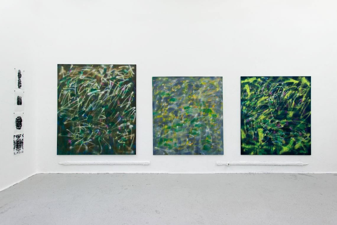 Foto: Ein Raum mit weißen Wänden und grauem Boden. An einer der Wände hängen drei großformatige Leinwände. An einer anderen Wand hängen mehre kleinformatige Papierarbeiten in einer Linie untereinander.