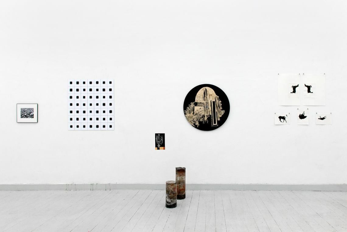 Foto: Ein Raum mit weißen Wänden und grauem Dielenboden. An den Wänden befinden sich mehrere flache Objekte, manche von ihnen sind quadratisch, eines ist rund. Vor der Wand stehen zwei Objekte im Raum.