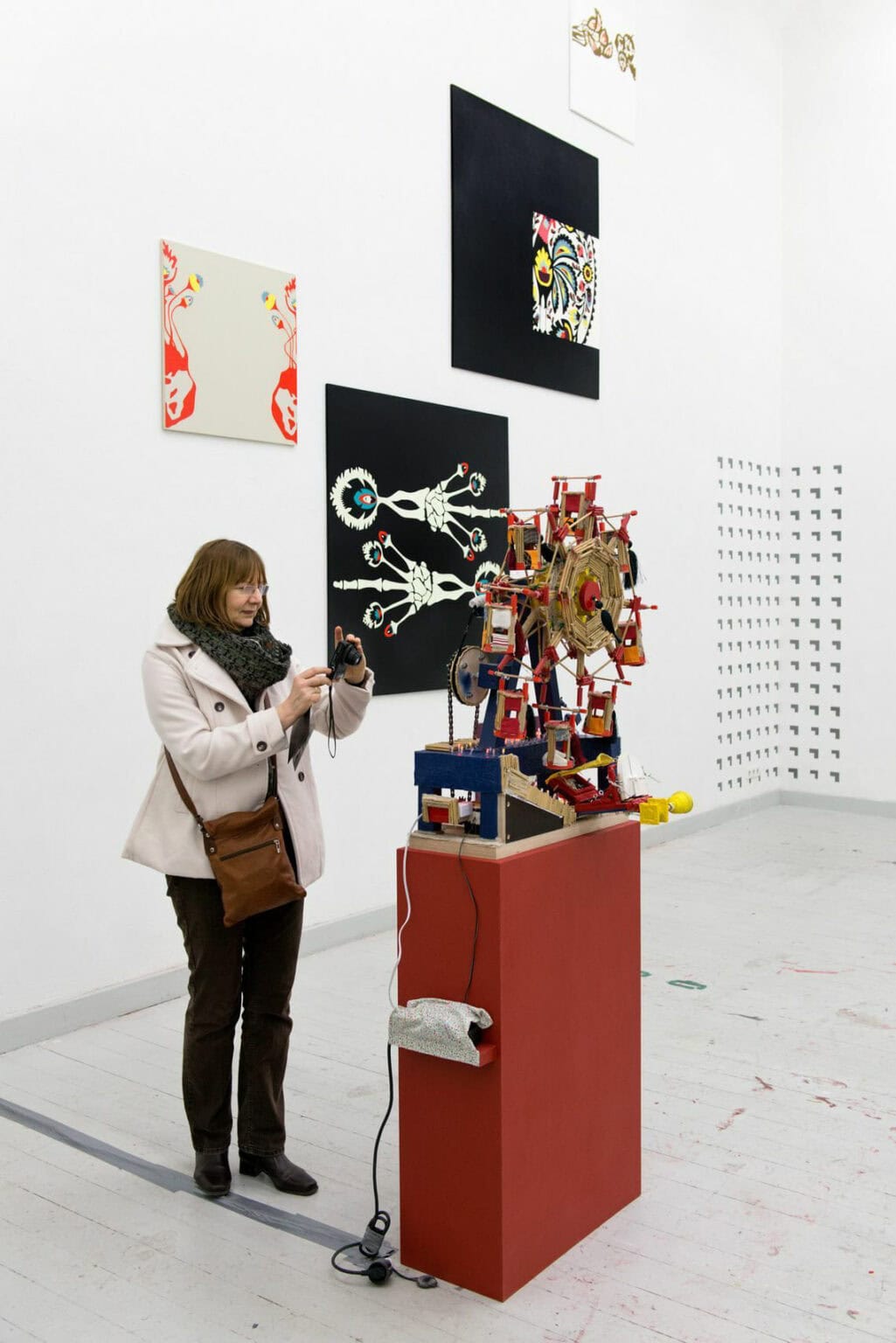 Foto: Ein Raum mit weißen Wänden und grauem Boden. An der Wand befinden sich mehre Leinwände, die auf unterschiedlicher höhe hängen. Teile der Wand sind mit einem sich wiederholenden Muster bemalt. Im Raum steht eine Skulptur auf einem Sockel. Im Raum steht eine person die eine Kamera in der Hand hält.