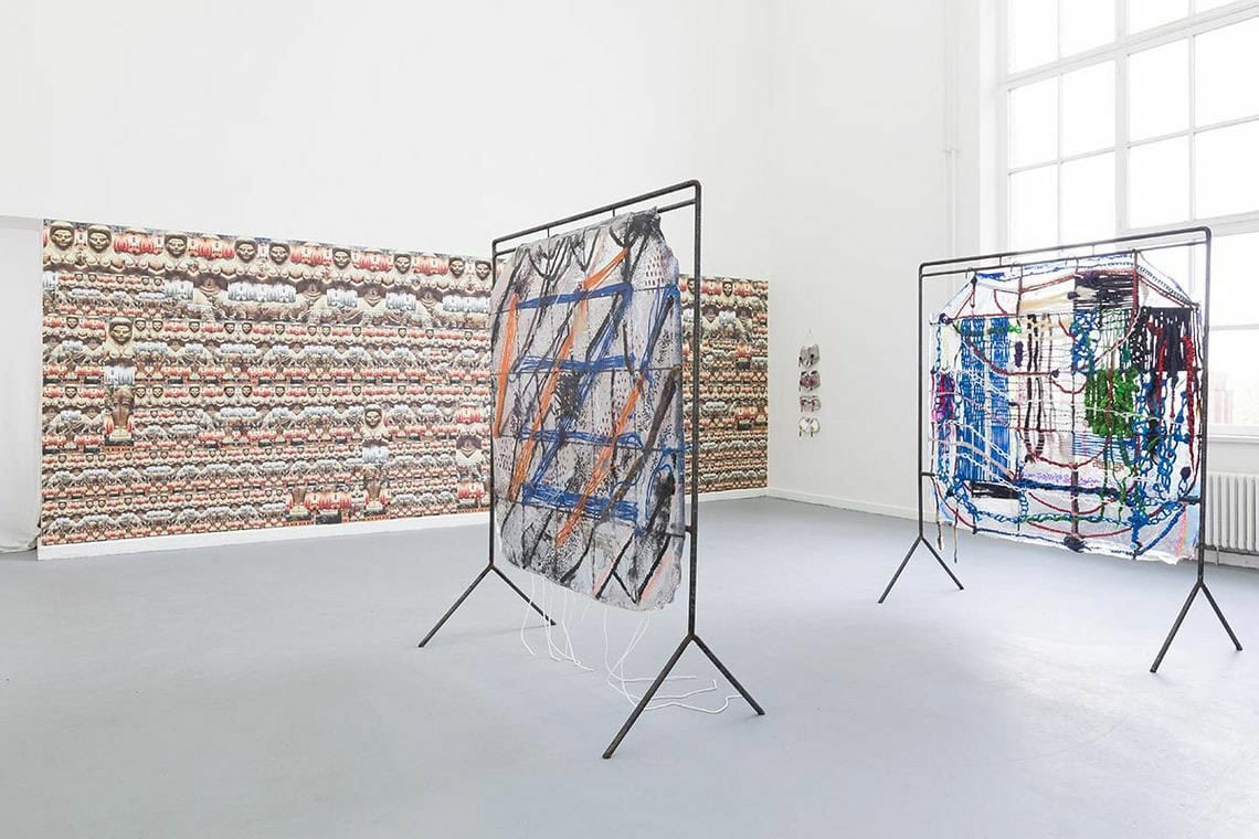 Foto: Ein Raum mit weißen Wänden, grauem Boden und großem Fenster. An einer der Wände befindet sich ein großformatiger, gemusterter Streifen. Im Raum stehen zwei flache Skulpturen.