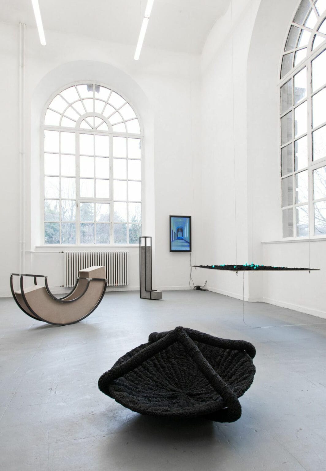 Foto: Ein Raum mit weißen Wänden, grauem Boden und großen Fenstern. Im Raum befinden sich mehrere große Skulpturen auf unterschiedlichen Materialien. Eine von Ihnen hängt von der Decke, die anderen liegen auf dem Boden. An der Wand hängt ein eingeschalteter Bildschirm.
