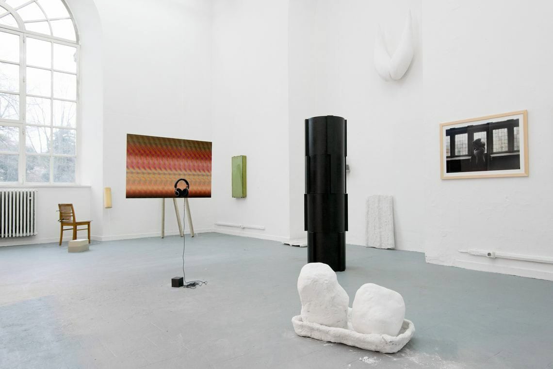 Foto: Ein Raum mit weißen Wänden, grauem Boden, einem großen Fenster und einer wandhohen, bogenförmigen Einbuchtung. An den Wänden befinden sich mehrere Objekte, einige von ihnen sind plastisch, andere flach. Sie hängen auf unterschiedlichen Höhen. Im Raum befinden sich mehrere Skulpturen, direkt auf dem Boden.