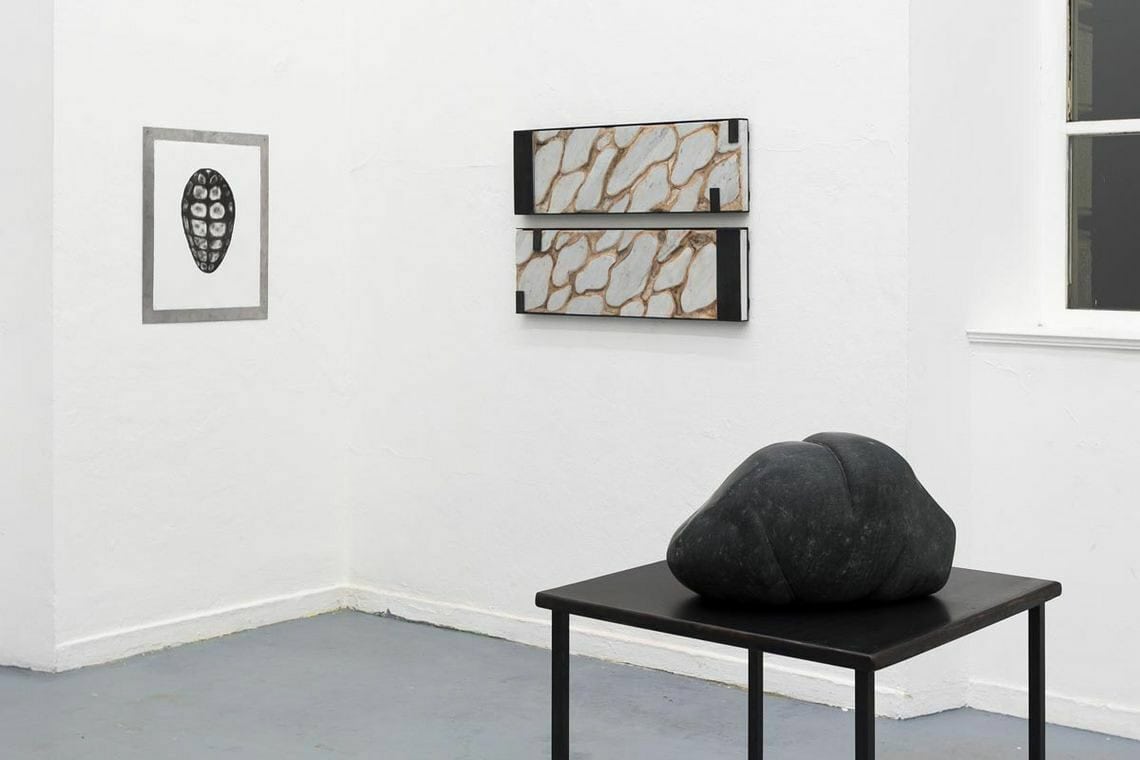 Foto: Ein Raum mit weißen Wänden und grauem Boden. An den Wänden hängen mehrere Flache Objekte. Im Raum steht eine Skulptur.