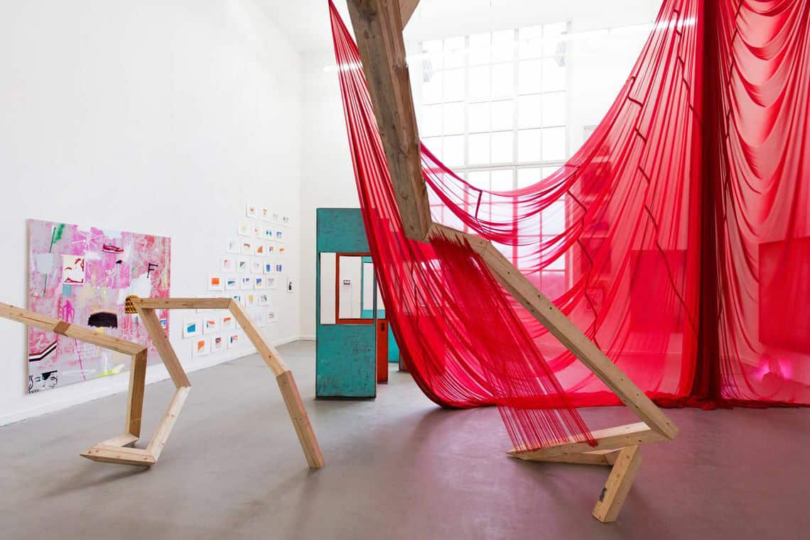 Foto: Ein Raum mit weißen Wänden, grauem Boden und einem großen Fenster. An den Wänden hängen mehrere flache Objekte neben- und untereinander. Im Raum befindet sich mehre große, zum Teil Raumhohe Skulpturen und Arrangements aus unterschiedlichen Materialien.