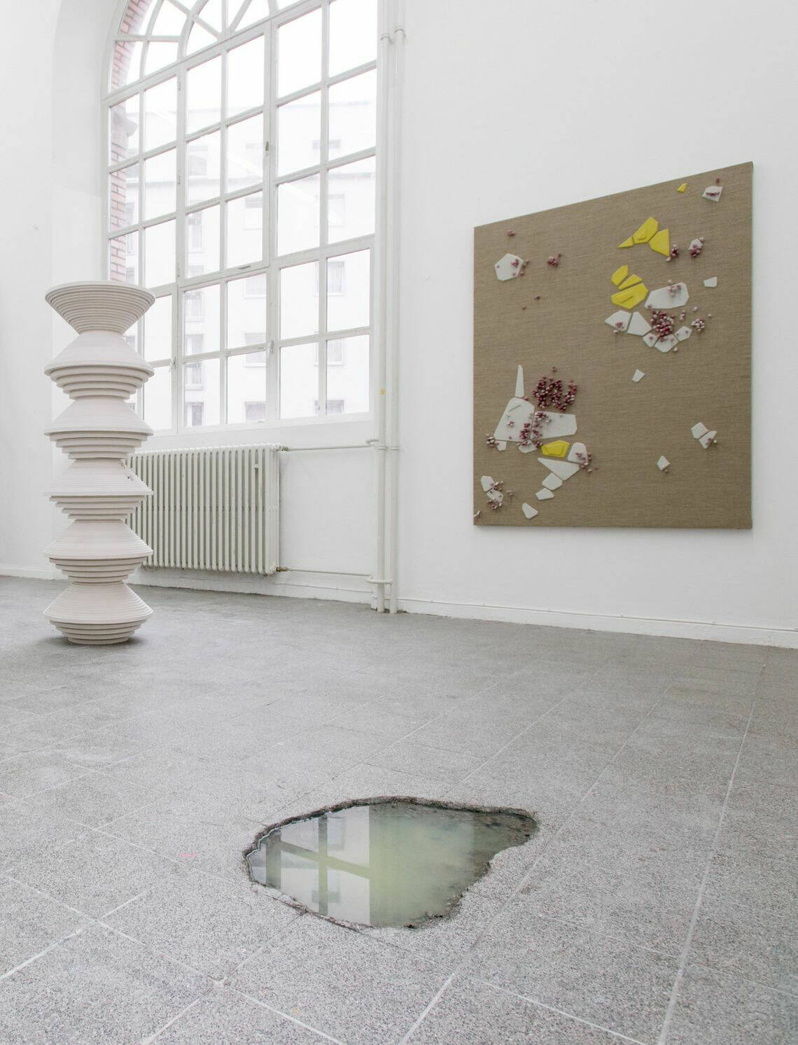 Foto: Ein Raum mit weißen Wänden, grauem, gefließten Steinboden und einem großen Fenster. An der Wand hängt eine großformatige Leinwand. Im Raum steht eine große Skulptur. In den Fliesen am Boden ist ein Loch, in dem sich trübes Wasser sammelt.