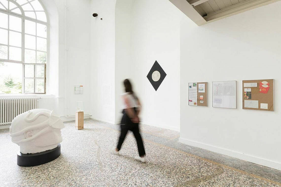 Foto: Ein Raum mit weißen Wänden, buntem Steinboden und einem großen Fenster. An den Wänden hängen klein- und mittelformatige, flache Objekte. Im Raum stehen zwei Skulpturen. Durch den Raum bewegt sich eine Person.