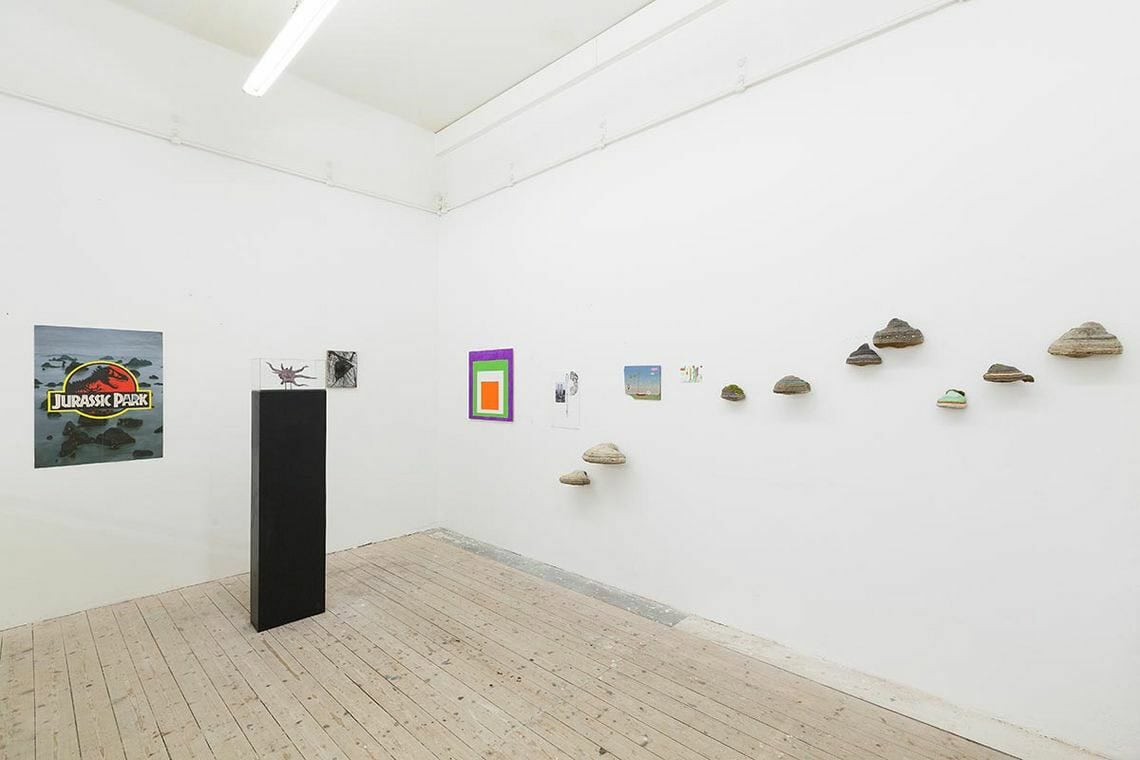 Foto: Ein Raum mit weißen Wänden und Dielenboden. Im Raum steht eine Skulptur. An den Wänden hängen, in einem Streifen angeordnet, viele klein- und mittelformatige Objekte.