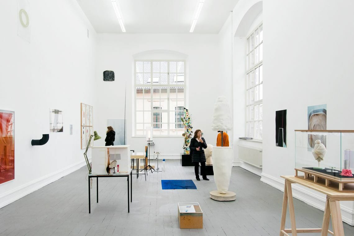 Ein Raum mit weißen Wänden, grauem Boden und großen Fenstern. An den Wänden befinden sich mehrere Objekte, einige von ihnen sind plastisch, andere flach. Sie hängen auf unterschiedlicher Höhe. Auf dem Boden liegen und stehen viele Objekte. Manche von ihnen sind zu mehreren auf flächigen Sockeln arrangiert, andere direkt auf dem Boden angebracht. Die Elemente stehen zum Teil sehr eng beieinander. Hinten im Raum befindet sich eine Person.