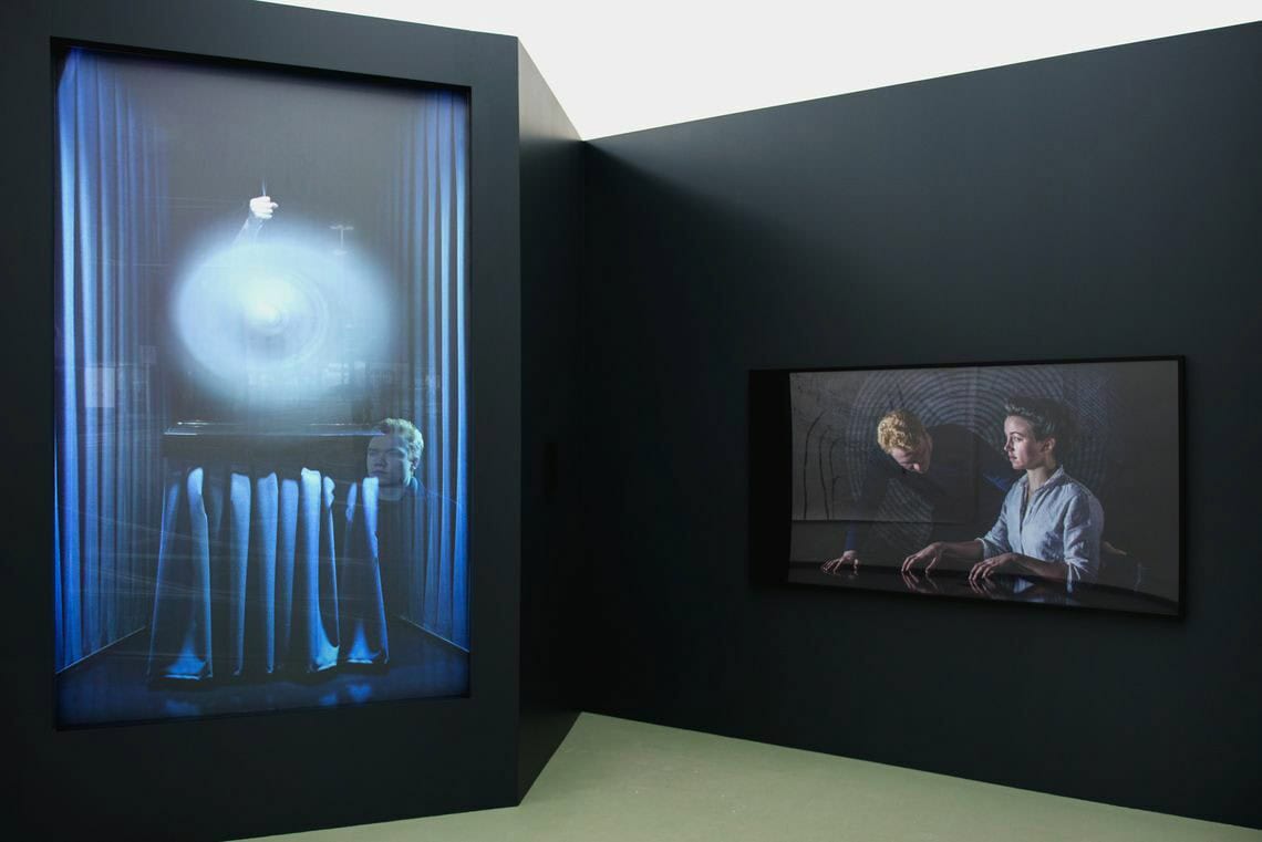 Foto: Ein Raum mit schwarzen Wänden und grauem Boden. Teile der Wand ergeben eine eckige Verkofferung. In dieser Verkofferung ist ein eingeschalteter Bildschirm eingelassen. Rechts daneben hängt ein weitere Bildschirm an der Wand.