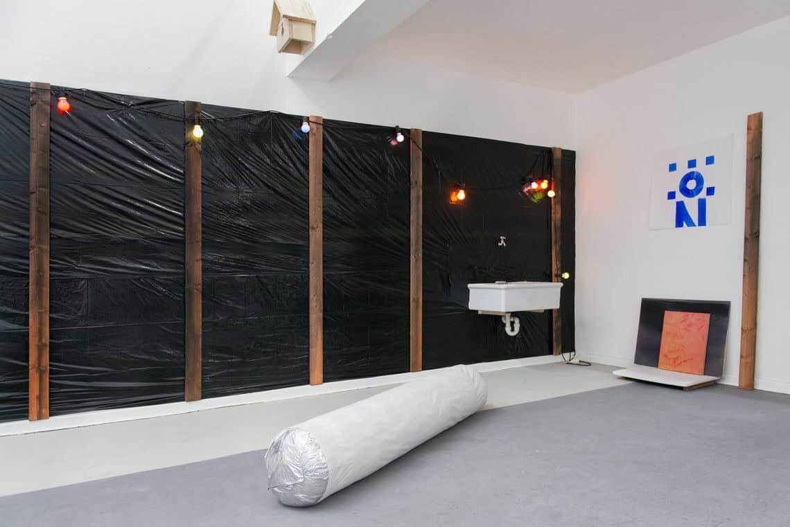 Foto: Ein Raum mit weißen Wänden und grauem Boden. Eine der Wände ist mit einer Konstruktion aus schwarzer Folie, Holzlatten und einer Lichterkette bedeckt. An den Wänden hängen und lehnen Objekte. Ein Streifen nahe der mit Folie bedeckten Wand ist hellgrau gestrichen, der Rest des Bodens dunkelgrau. Im Raum liegt ein großes, langes Objekt auf dem Boden.