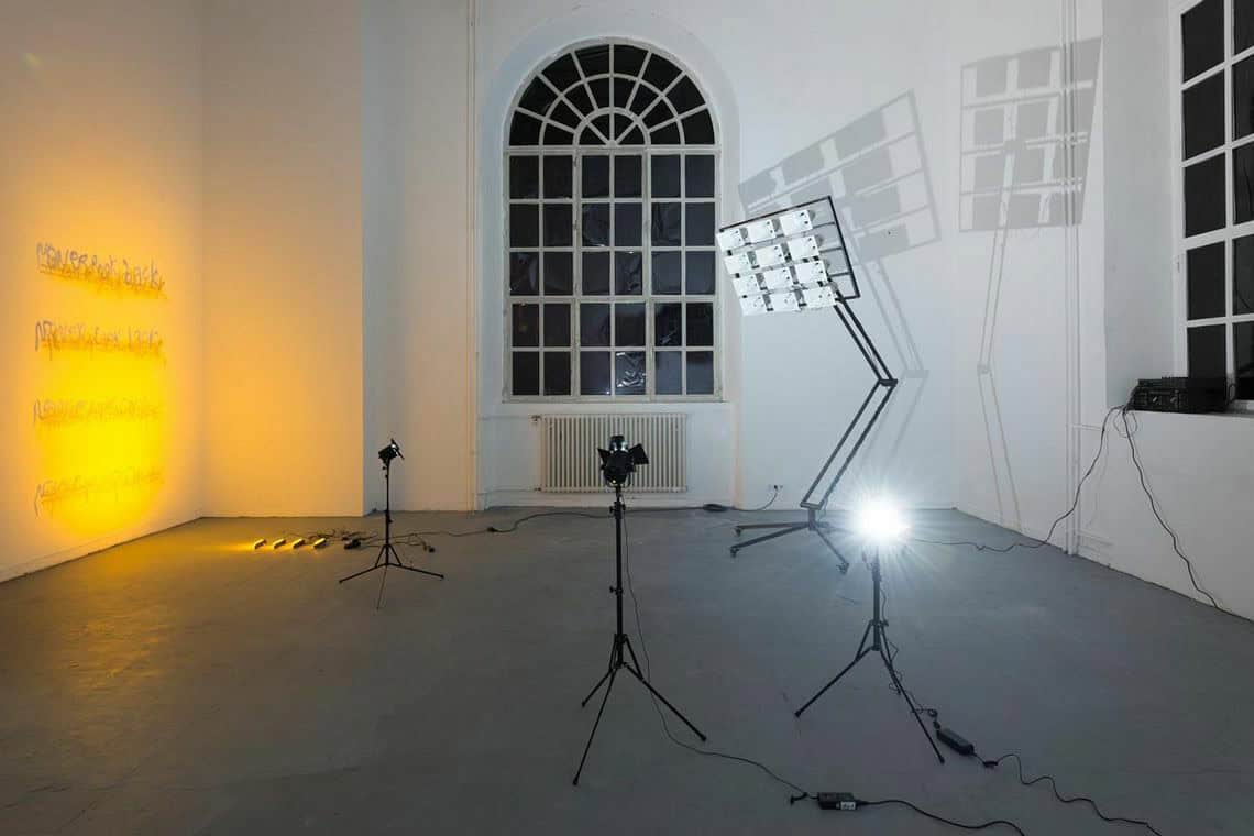 Foto: Ein Raum mit weißen Wänden, grauem Boden und zwei großen Bogenfenstern. An einer der Wände befinden sich Objekte, die von einem hellen, warmen, gelben Licht angestrahlt werden, das von auf dem Boden liegenden Lichtquellen ausgeht. Im Raum verteilt befinden sich Lichter auf Stativen und eine Konstruktion auf Rollen, an deren Kopf sich eine Gruppe von Lautsprechern bzw. Sirenen befindet. Die Lichter strahlen das Wandobjekt und die Lautsprecher Konstruktion an. Letztere wirft zwei große Schatten.