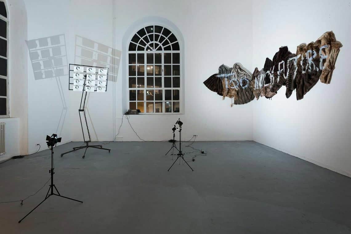 Foto: Ein Raum mit weißen Wänden, grauem Boden und zwei großen Bogenfenstern. Oben an der Wand befindet sich eine Gruppe von Objekten, auf denen "TOO MUCH FUTURE" steht. Im Raum verteilt befinden sich Lichter auf Stativen und eine Konstruktion auf Rollen, an deren Kopf sich eine Gruppe von Lautsprechern bzw. Sirenen befindet. Die Lichter strahlen das Wandobjekt und die Lautsprecher Konstruktion an. Letztere wirft zwei große Schatten.