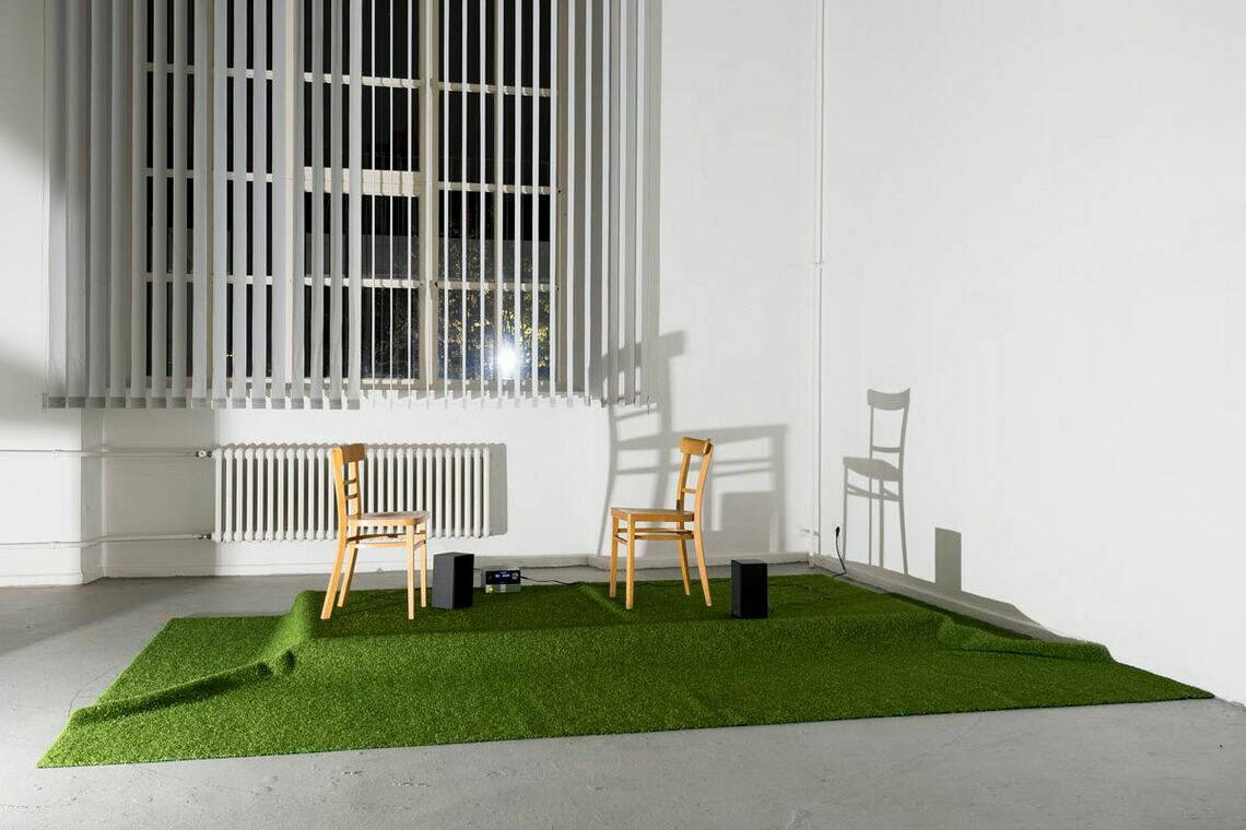 Foto: Ein abgedunkelter Raum mit weißen Wänden, grauem Boden und einem großen Fenster, vor dem Lamellen-Vorhänge hängen. Teile des Bodens sind mit Kunstrasen bedeckt, unter dem sich ein Podest abzeichnet. Auf dem Podest stehen zwei Stühle, zwei Leitsprecher und Technik. Die Szenerie wird von einem Licht angestrahlt, die Objekte werfen Schatten.