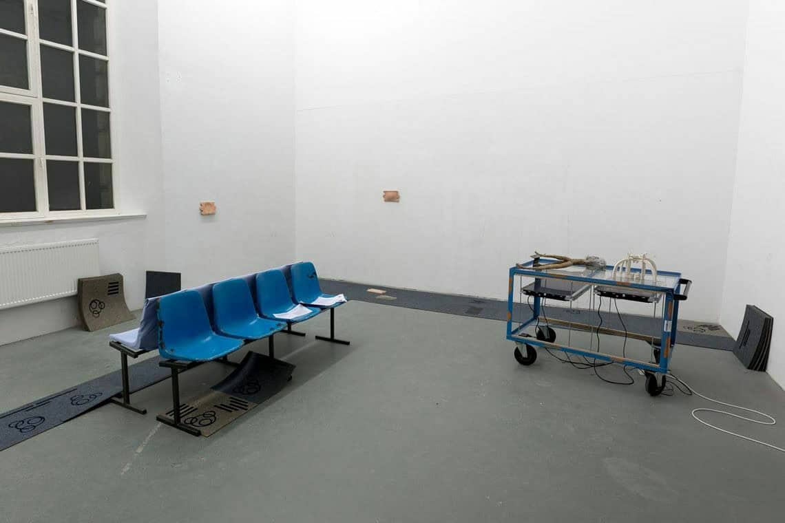 Foto: Ein Raum mit weißen Wänden, grauem Boden und einem großen Fenster. An der Wand des Raumes befinden sich zwei kleine Objekte. Im Raum liegen Streifen aus Teppich. Weitere quadratische Teppichstücke lehnen an der Wand. Im Raum stehen zwei Sitzbänke mit Schalenstühlen, rücken an Rücken aneinander. Im Raum steht außerdem ein Rollwagen, auf dem sich Objekte und Kabel befinden.