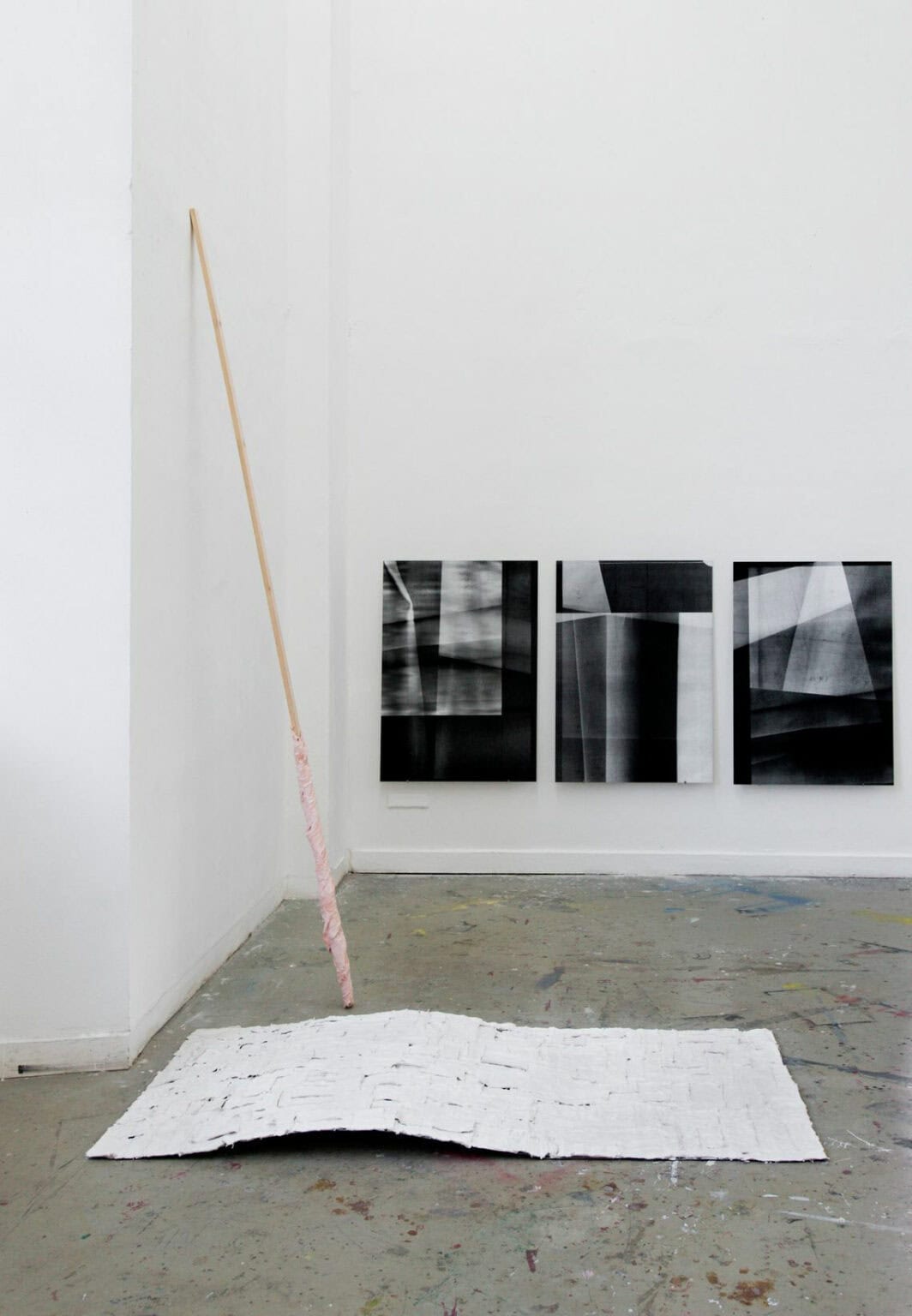 Foto: Ein Raum mit weißen Wänden und grauem Boden. Der Boden ist mit Farbsprenklern übersehen. Auf dem Boden liegt und an der Wand lehnt ein Objekt. An der Wand hängen drei flache Objekte im gleichem Format im gleichen Abstand zueinander.