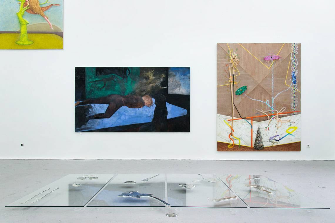 Foto: Ein Raum mit weißen Wänden und grauem Boden. An der Wand des Raumes hängen Leinwände im Großformat. Auf dem Boden befinden sich drei unterschiedlich große Glasscheiben, auf denen flache Objekte liegen.