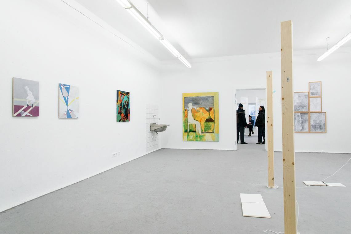 Foto: Ein Raum mit weißen Wänden und grauem Boden An der Wand des Raumes hängen zwei Leinwände in Klein- und Großformat sowie gerahmte Objekte. Im Raum stehen Holzlatten, an denen schüre befestigt sind. Auf dem Boden liegen aufgeschlagene Hefte.