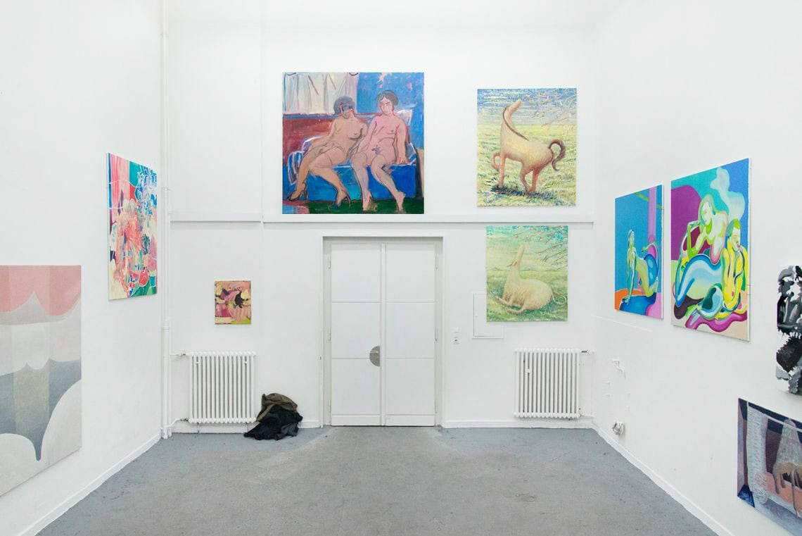 Foto: Ein Raum mit weißen Wänden und grauem Boden. An den Wänden hängen mehrere Leinwände in Klein-, Mittel- und Großformaten neben und übereinander.