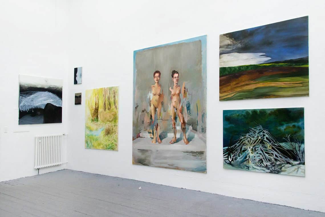 Foto: Ein Raum mit weißen Wänden und grauem Dielenboden. An den Wänden hängen mehrere Leinwände in Klein-, Mittel- und Großformaten neben und übereinander.