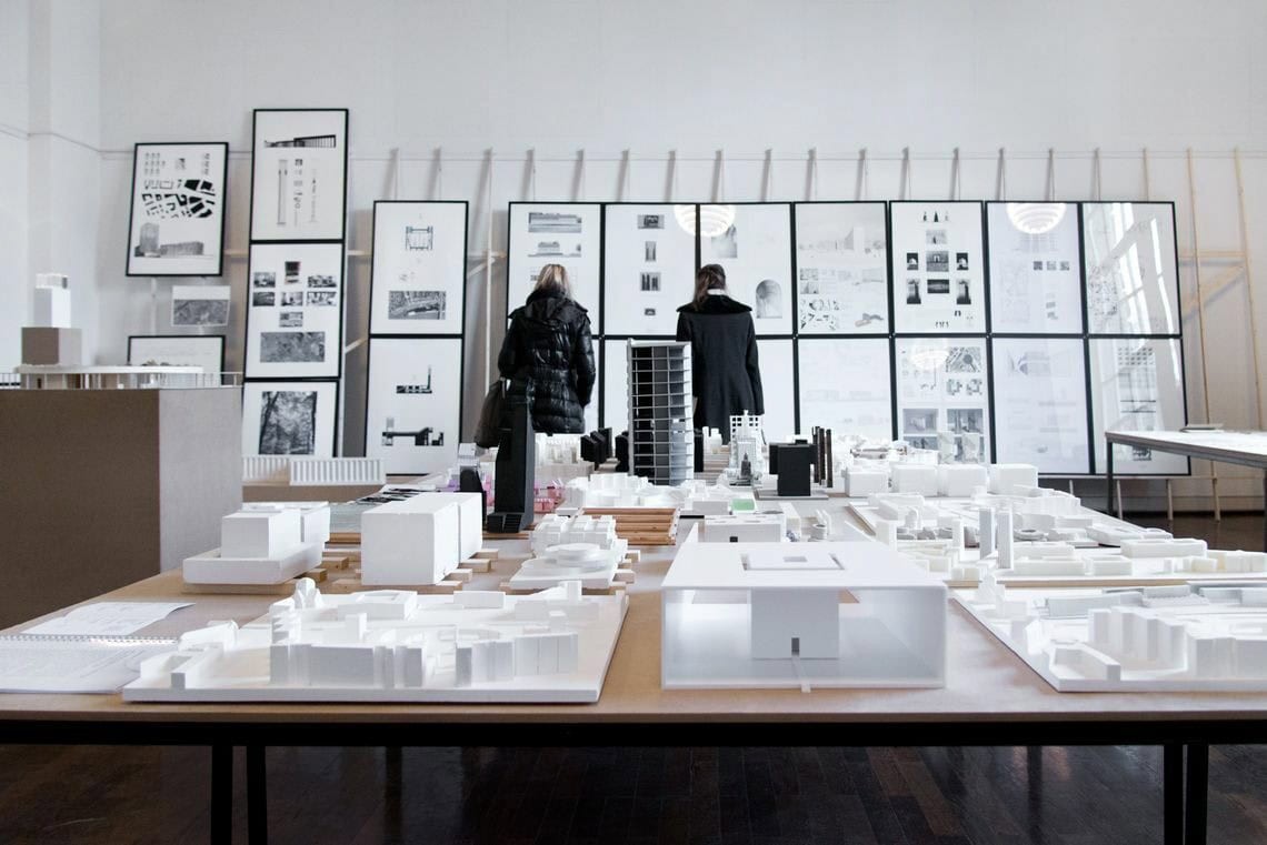 Foto: Ein Raum mit weißen Wänden und braunem Dielenboden. Im Raum stehen mehrere Tische, auf denen sich modellartige Objekte befinden. Hinten an der Wand des Raumes lehnt eine Konstruktion aus Holzlatten. Auf die Holzlatten sind zwei Reihen untereinander gerahmte Objekte angebracht, die alle das gleiche Format haben. Die Konstruktion bedeckt die gesamte länge der Wand.