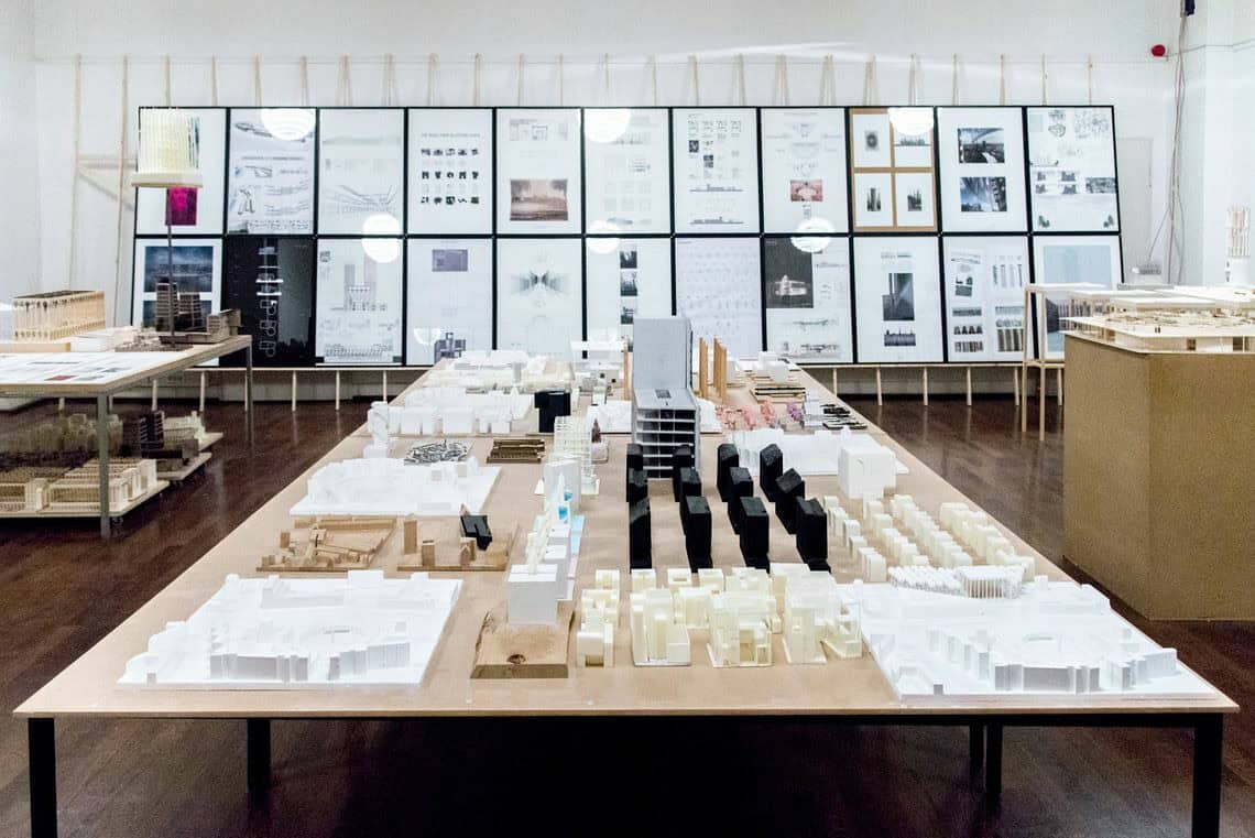 Foto: Ein Raum mit weißen Wänden und braunem Dielenboden. Im Raum stehen mehrere Tische, auf denen sich modellartige Objekte befinden. Hinten an der Wand des Raumes lehnt eine Konstruktion aus Holzlatten. Auf die Holzlatten sind zwei Reihen untereinander gerahmte Objekte angebracht, die alle das gleiche Format haben. Die Konstruktion bedeckt die gesamte länge der Wand.